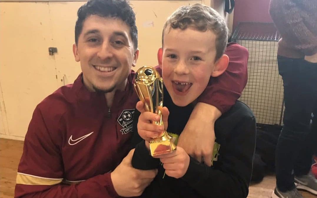 A First Steps Soccer coach hugging a young player holding a trophy