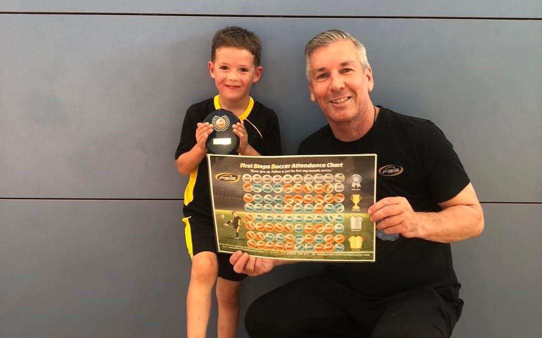 A young First Steps Soccer player with an award and their coach holding the attendance chart