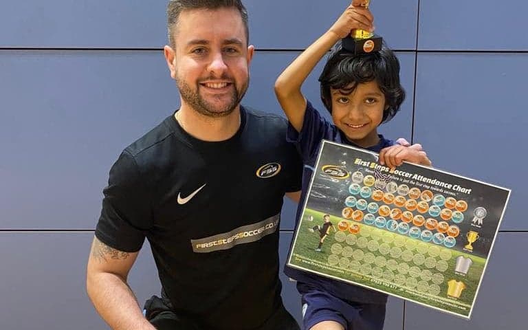 A young First Steps Soccer player holding a trophy and attendance chart with their coach